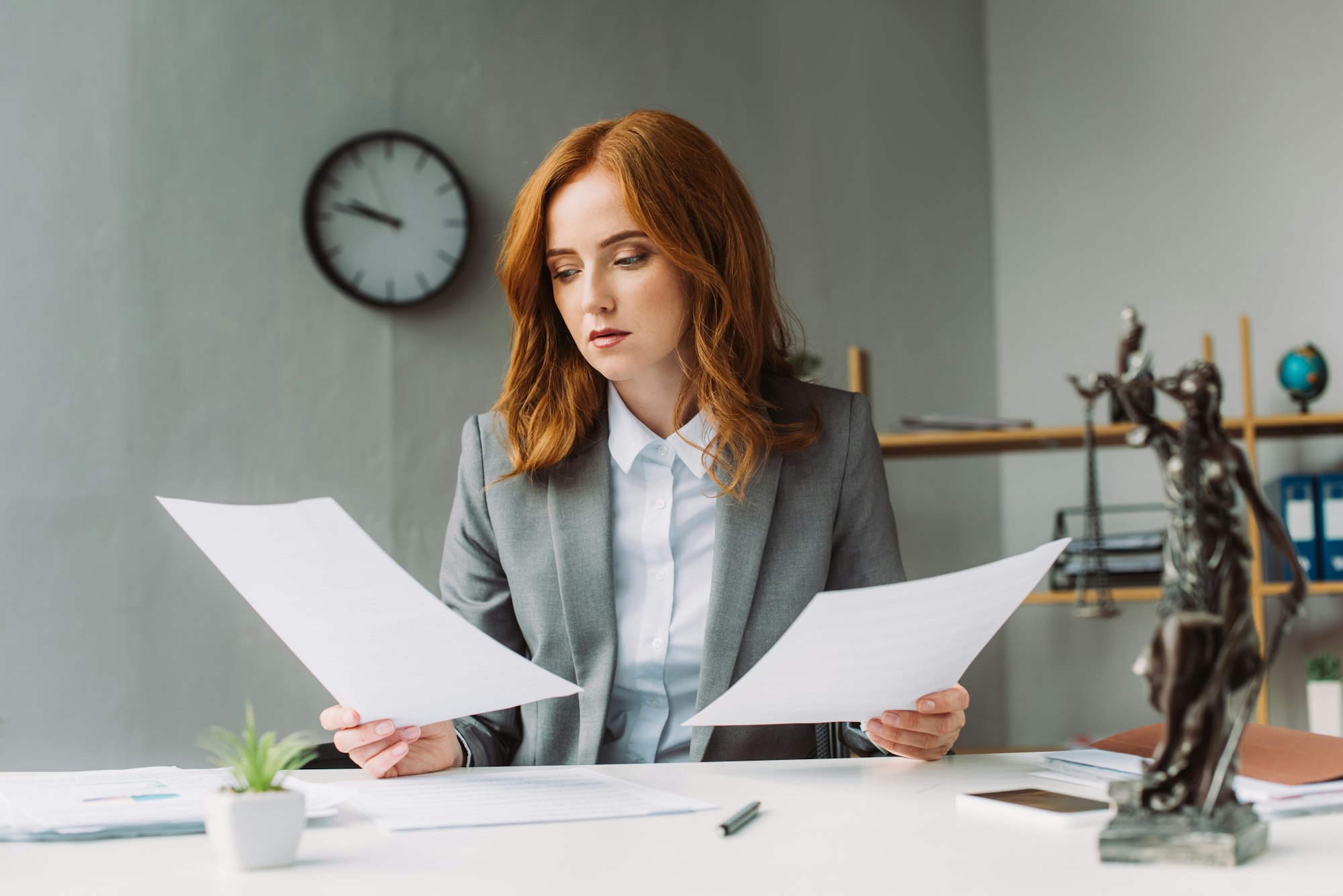 Redhead female lawyer looking at paper sheet, while sitting at workplace with blurred themis